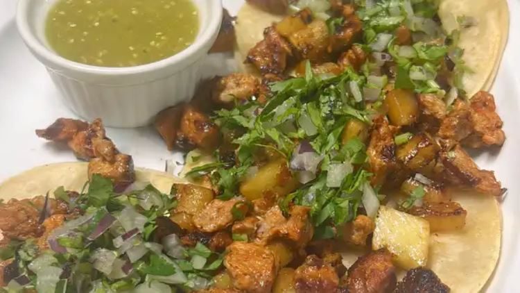Open faced al pastor tacos with charred pork, grilled pineapple, cilantro and onion beside a ramekin of green salsa.
