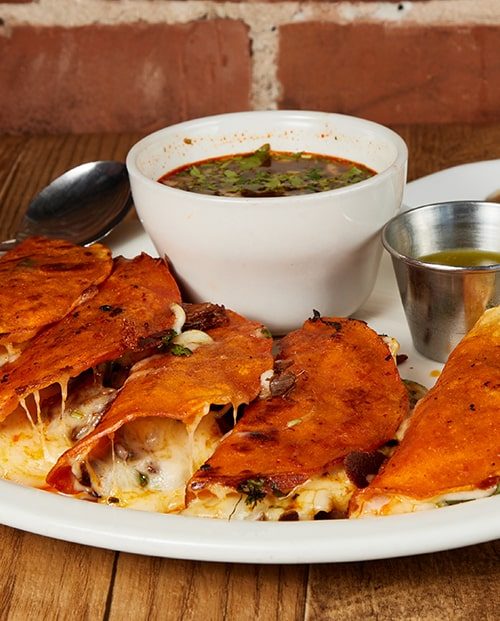 Five crisped birria quesa tacos on a white plate beside a bowl of dark consomé and a cup of green salsa.