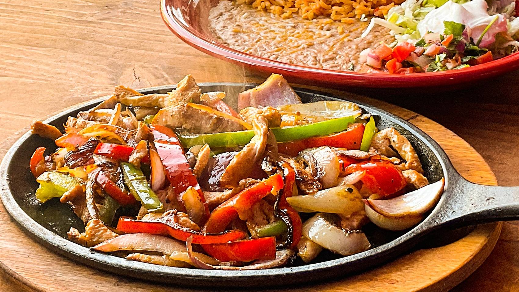 A cast iron skillet of chicken fajitas with peppers and onions still steaming, beside a plate of refried beans, Mexican rice and pico de gallo.